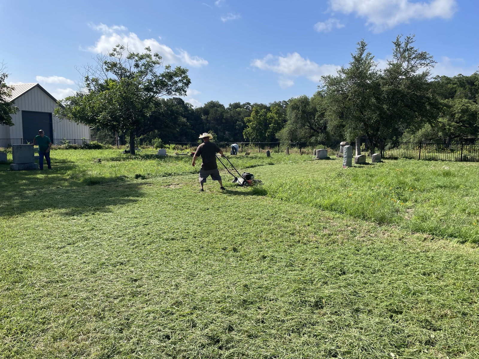 A volunteer pushing a mower cuts a wide swath through tall grass at the cemetery, headstones and the equipment shed in the background.