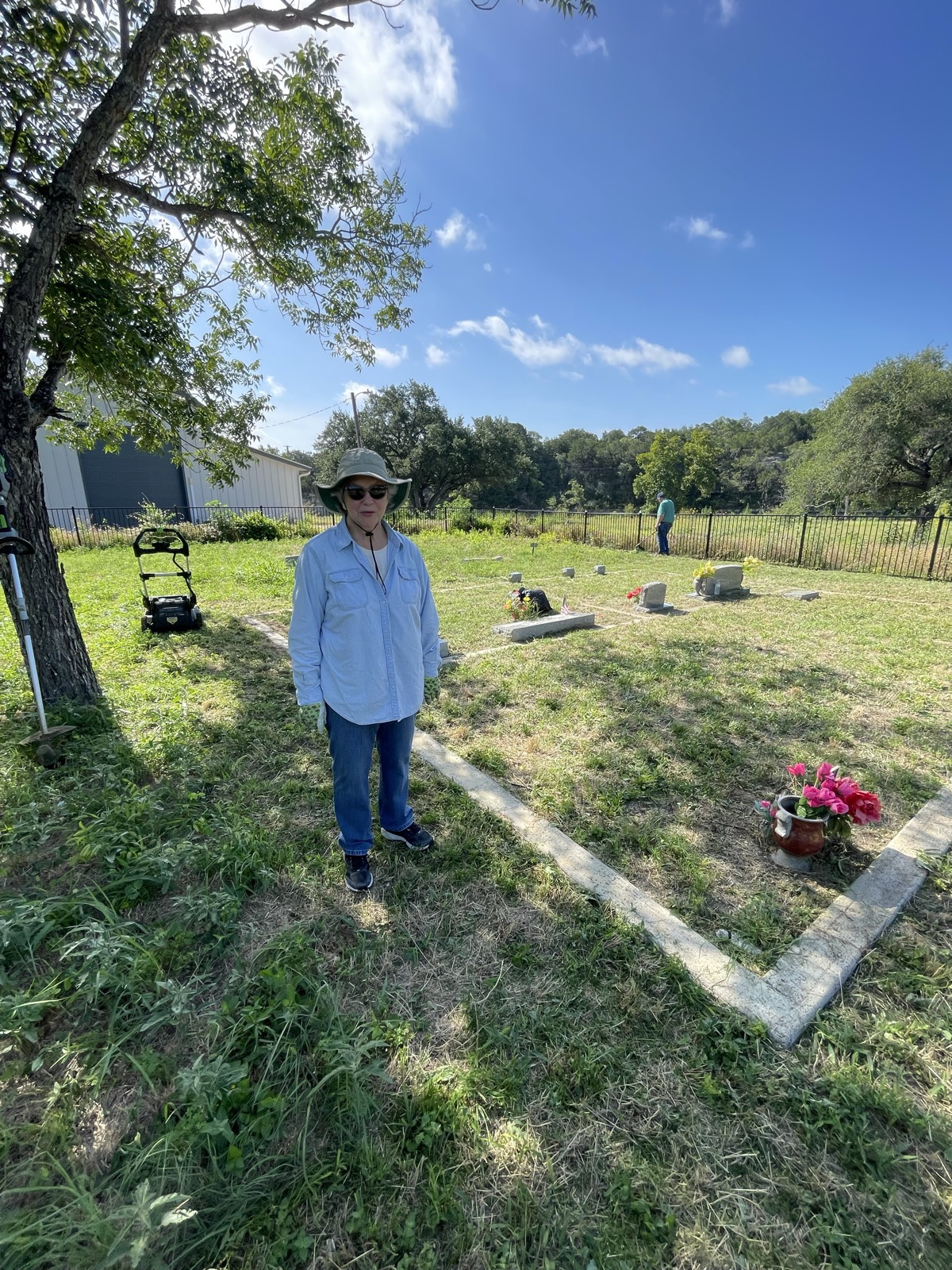 A volunteer in a sun hat stands beside a curbed family plot with pink flowers, a mower parked under a tree nearby.
